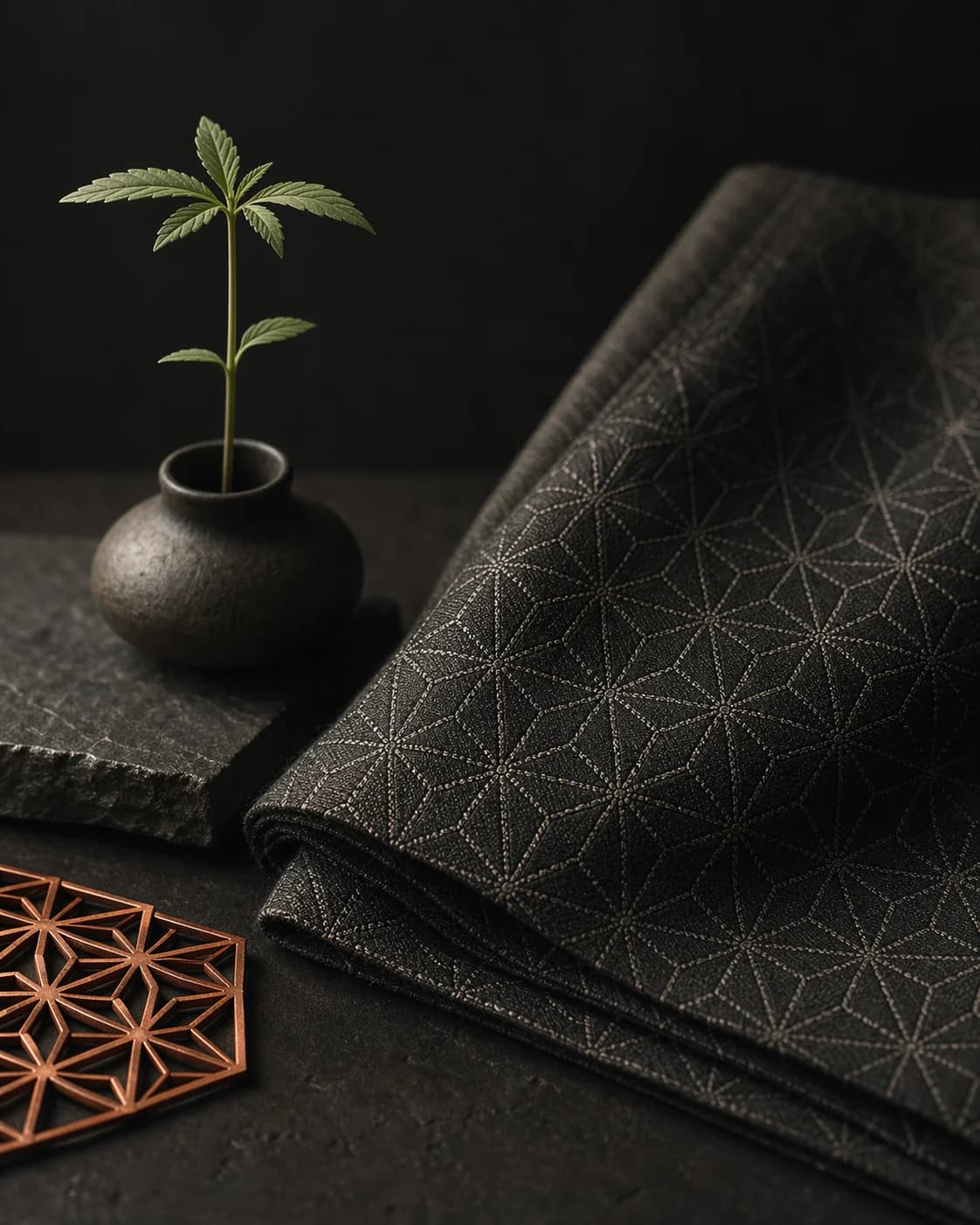 A folded asanoha-patterned textile beside a small plant in a dark editorial still life.